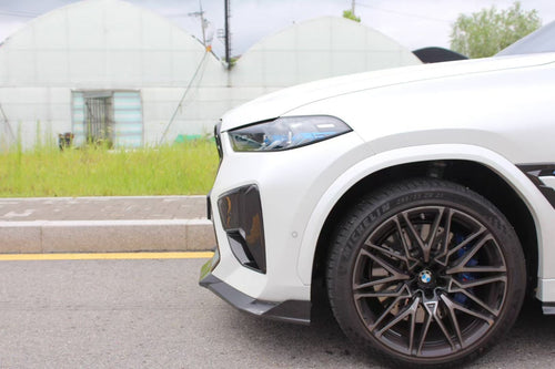 Front view of a white BMW X6M featuring an aggressive REVOZPORT front bumper and carbon fiber aero styling parked on a quiet urban street.