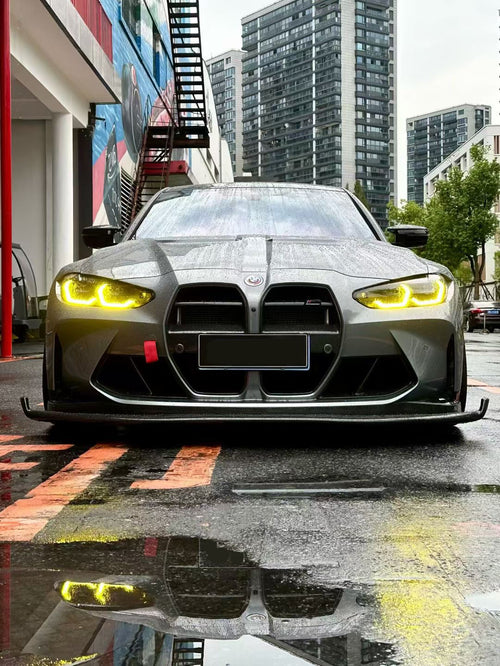 Gray BMW G80 M3 with aggressive carbon fiber aero kit, front splitter, side skirts, and rear wing parked on a rainy urban street with yellow DRL headlights illuminated.