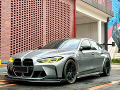 Gray BMW G80 M3 with aggressive carbon fiber aero kit, front splitter, side skirts, and rear wing parked on a rainy urban street with yellow DRL headlights illuminated.