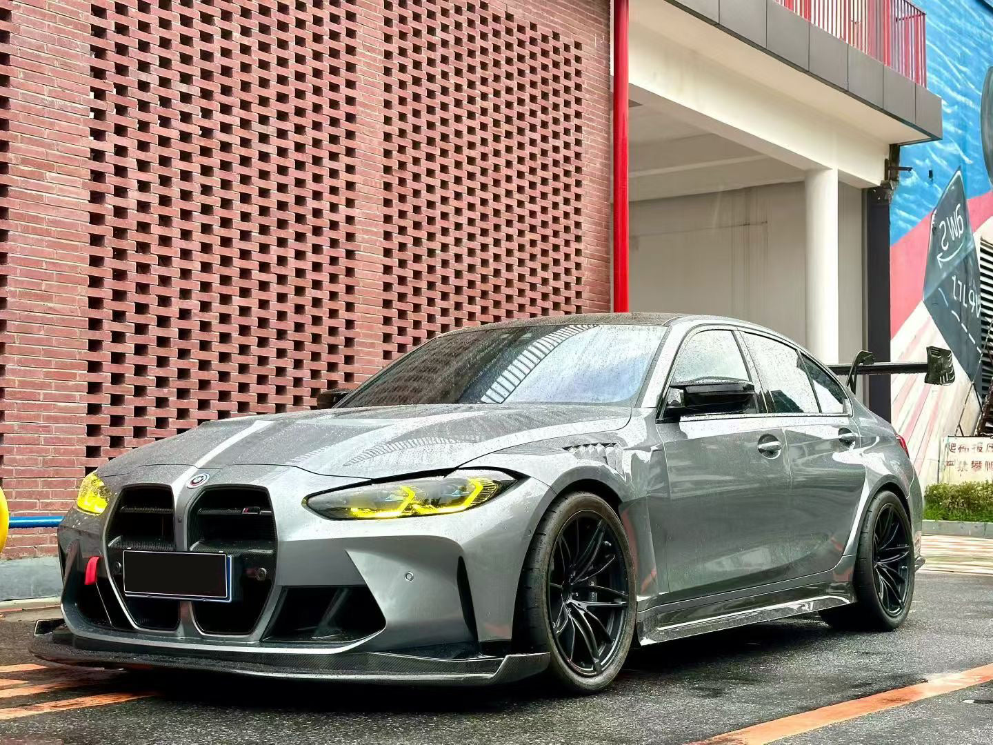 Gray BMW G80 M3 with aggressive carbon fiber aero kit, front splitter, side skirts, and rear wing parked on a rainy urban street with yellow DRL headlights illuminated.