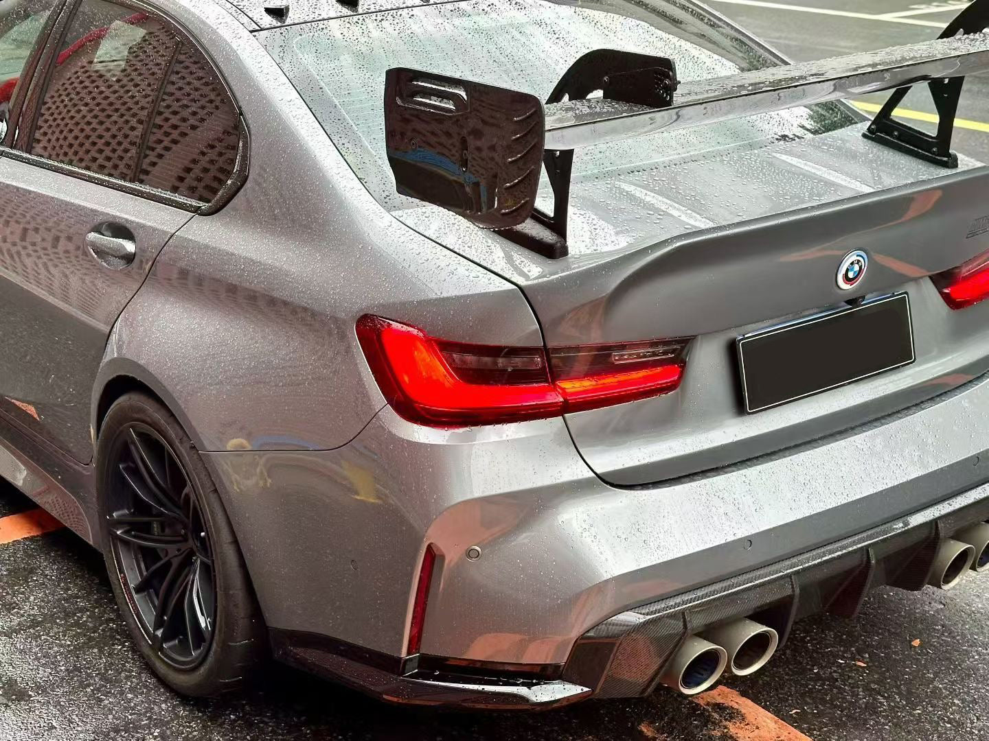 Gray BMW G80 M3 with aggressive carbon fiber aero kit, front splitter, side skirts, and rear wing parked on a rainy urban street with yellow DRL headlights illuminated.