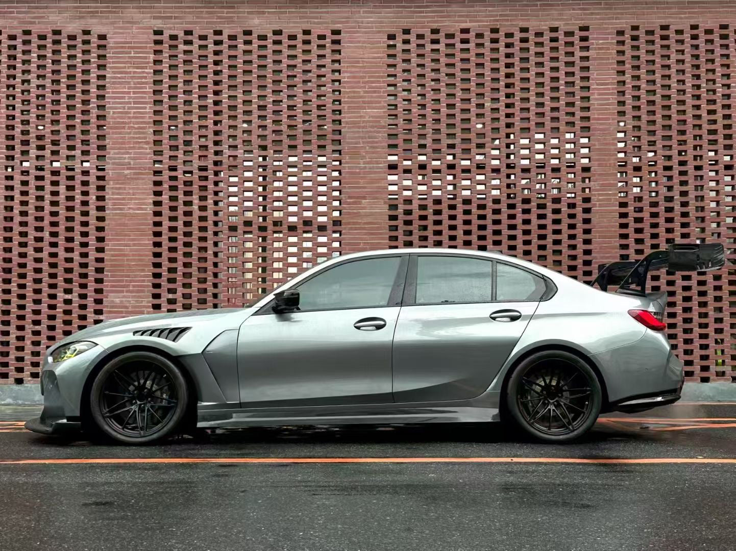 Gray BMW G80 M3 with aggressive carbon fiber aero kit, front splitter, side skirts, and rear wing parked on a rainy urban street with yellow DRL headlights illuminated.