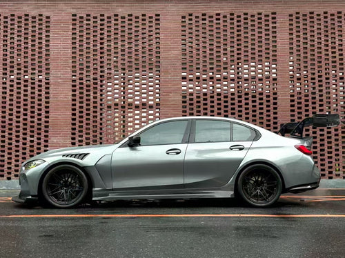 Gray BMW G80 M3 with aggressive carbon fiber aero kit, front splitter, side skirts, and rear wing parked on a rainy urban street with yellow DRL headlights illuminated.