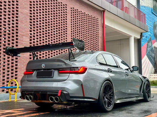 Gray BMW G80 M3 with aggressive carbon fiber aero kit, front splitter, side skirts, and rear wing parked on a rainy urban street with yellow DRL headlights illuminated.