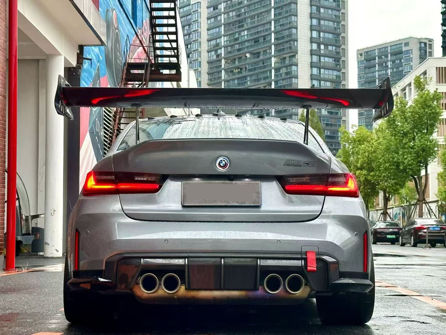 Gray BMW G80 M3 with aggressive carbon fiber aero kit, front splitter, side skirts, and rear wing parked on a rainy urban street with yellow DRL headlights illuminated.