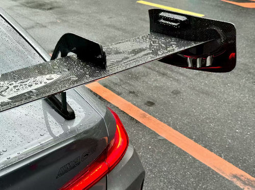 Gray BMW G80 M3 with aggressive carbon fiber aero kit, front splitter, side skirts, and rear wing parked on a rainy urban street with yellow DRL headlights illuminated.