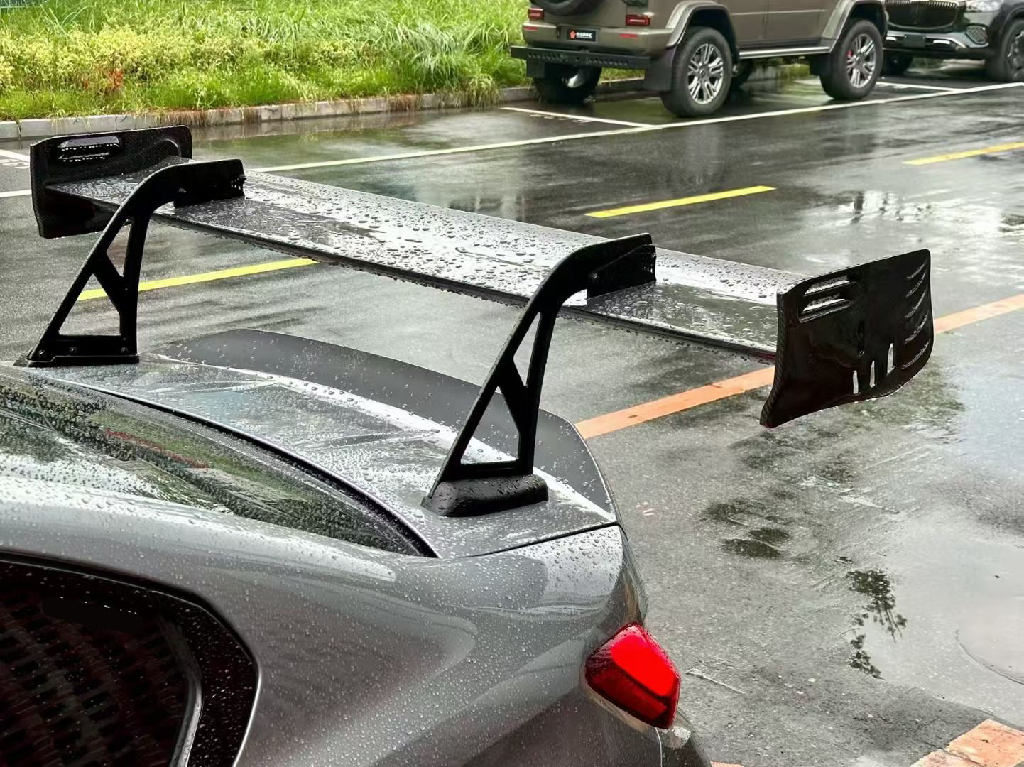 Gray BMW G80 M3 with aggressive carbon fiber aero kit, front splitter, side skirts, and rear wing parked on a rainy urban street with yellow DRL headlights illuminated.
