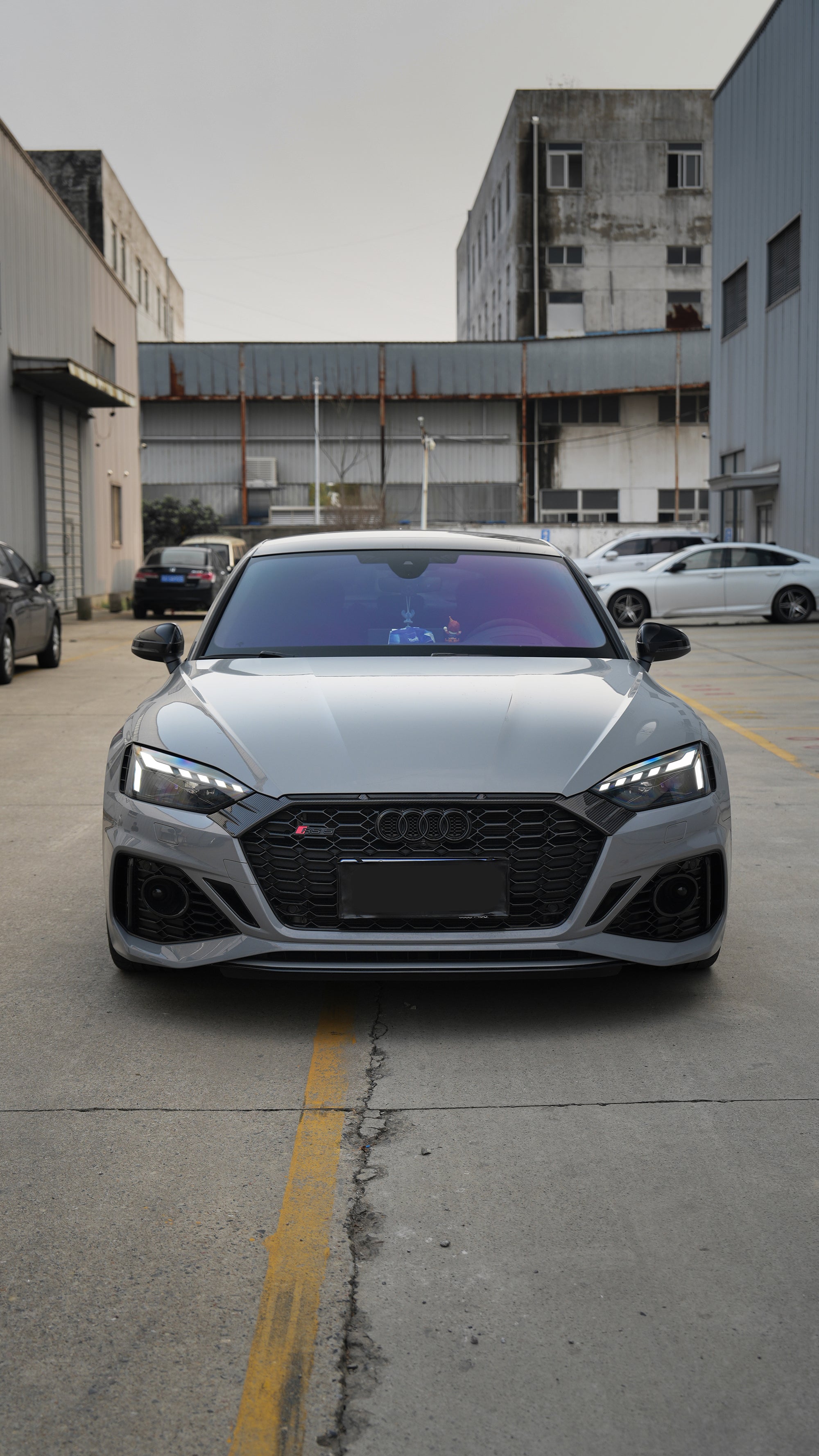 Front view of a gray Audi RS5 Sportback featuring a REVOZPORT carbon fiber body kit with aggressive front bumper styling and black honeycomb grille parked in an industrial lot.