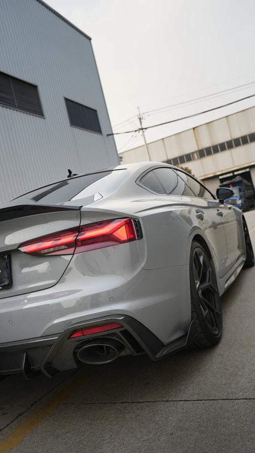 Front view of a gray Audi RS5 Sportback featuring a REVOZPORT carbon fiber body kit with aggressive front bumper styling and black honeycomb grille parked in an industrial lot.