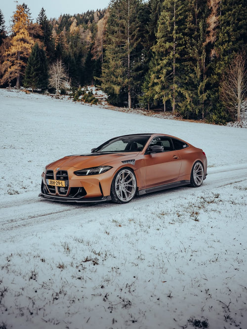 Close-up of REVOZPORT forged multi-spoke wheels mounted on a bronze widebody sports car with carbon fiber aero details on a snowy road.
