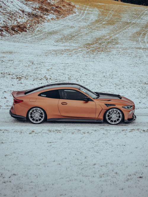 Close-up of REVOZPORT forged multi-spoke wheels mounted on a bronze widebody sports car with carbon fiber aero details on a snowy road.