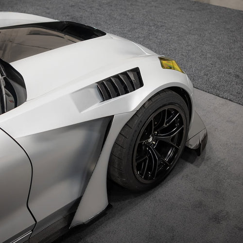 Close-up of a silver performance car’s front wheel and brake setup, featuring black forged multi-spoke wheels, large drilled brake rotors, and high-performance racing tires. The image also highlights the aggressive wide-body fender design and carbon fiber aerodynamic components in a motorsport environment.