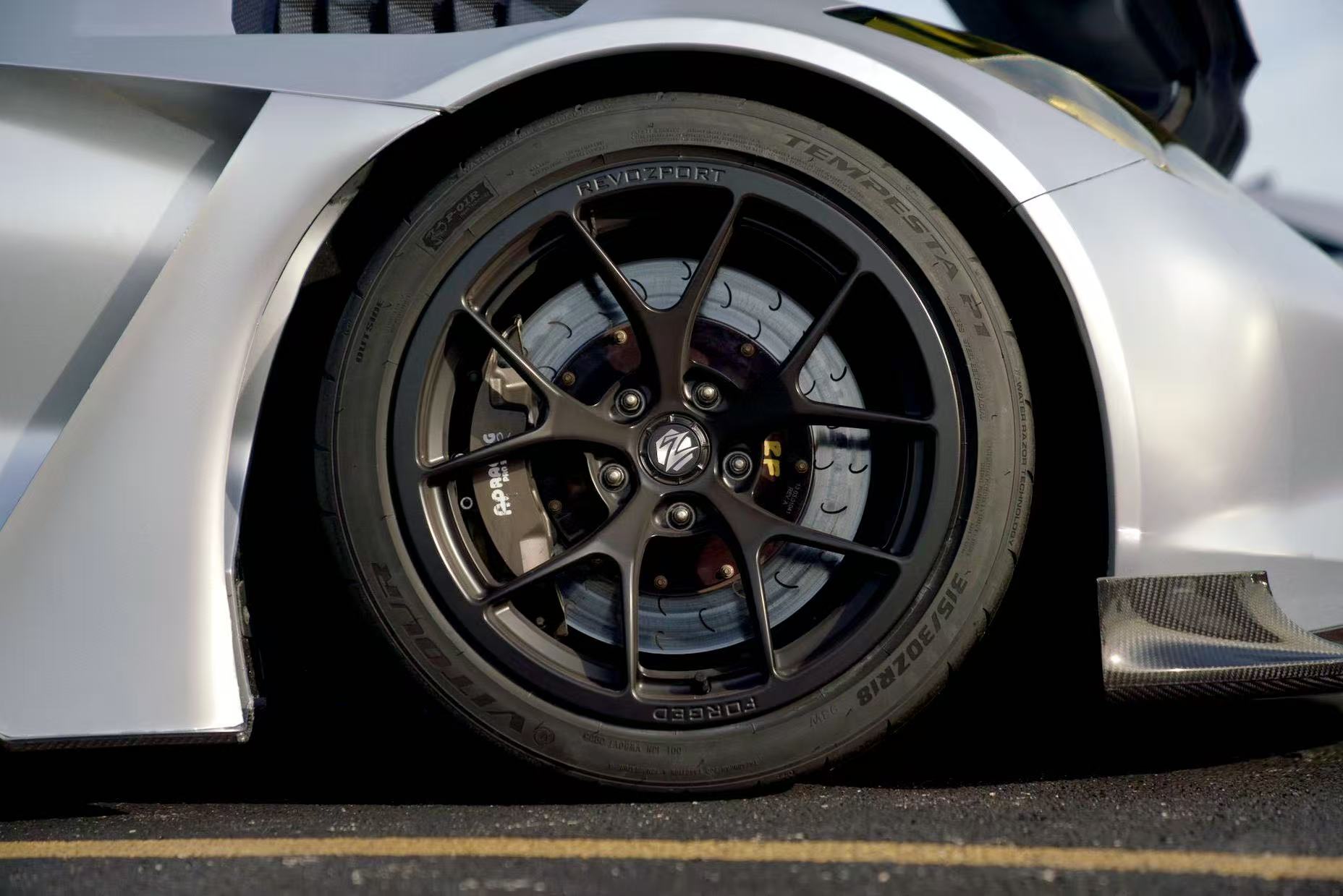 Close-up of a silver performance car’s front wheel and brake setup, featuring black forged multi-spoke wheels, large drilled brake rotors, and high-performance racing tires. The image also highlights the aggressive wide-body fender design and carbon fiber aerodynamic components in a motorsport environment.