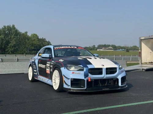 A light blue and white BMW race car parked in a racetrack paddock, featuring a wide-body aerodynamic kit, carbon fiber front splitter, large air intakes, and white racing wheels. The car is covered with sponsor decals and track graphics, with racetrack barriers and trees visible in the background.
