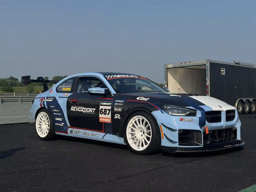 A light blue and white BMW race car parked in a racetrack paddock, featuring a wide-body aerodynamic kit, carbon fiber front splitter, large air intakes, and white racing wheels. The car is covered with sponsor decals and track graphics, with racetrack barriers and trees visible in the background.
