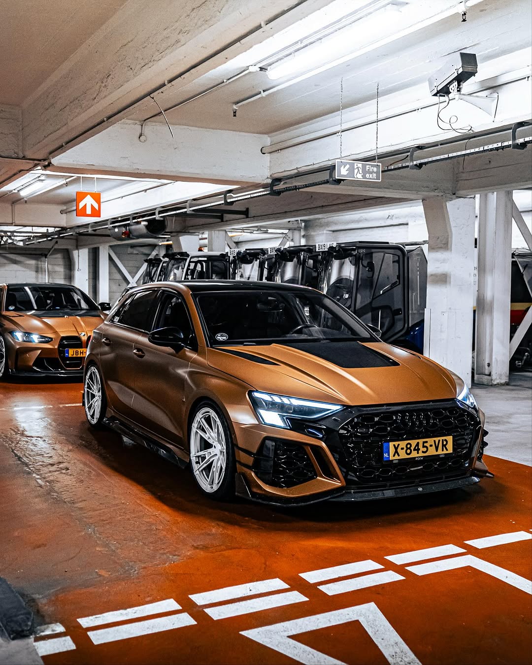 Bronze BMW G82 M4 fitted with REVOZPORT forged silver multi-spoke wheels, parked on a luxury city street alongside a matching Audi RS model.