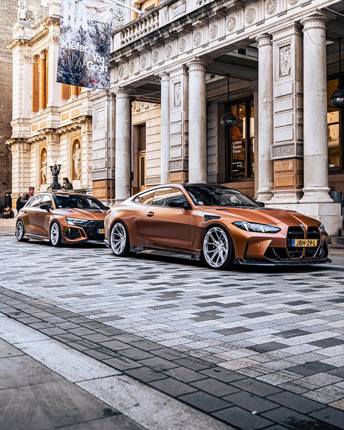 Bronze BMW G82 M4 fitted with REVOZPORT forged silver multi-spoke wheels, parked on a luxury city street alongside a matching Audi RS model.