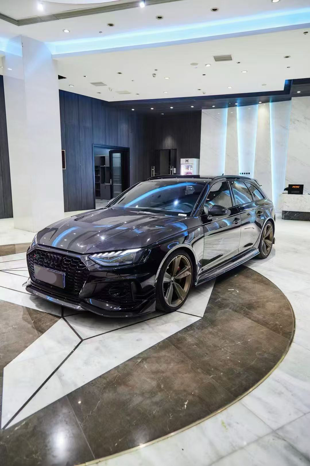 Black Audi RS4 Avant featuring a REVOZPORT carbon fiber body kit with front lip, side skirts, and aggressive aero styling displayed inside a luxury showroom.