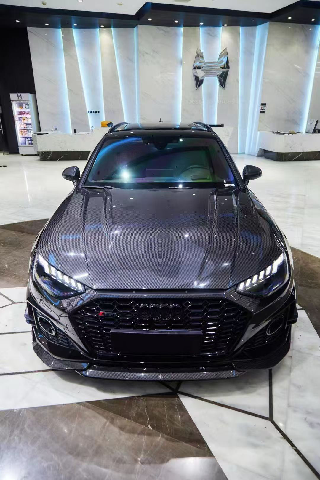 Black Audi RS4 Avant featuring a REVOZPORT carbon fiber body kit with front lip, side skirts, and aggressive aero styling displayed inside a luxury showroom.
