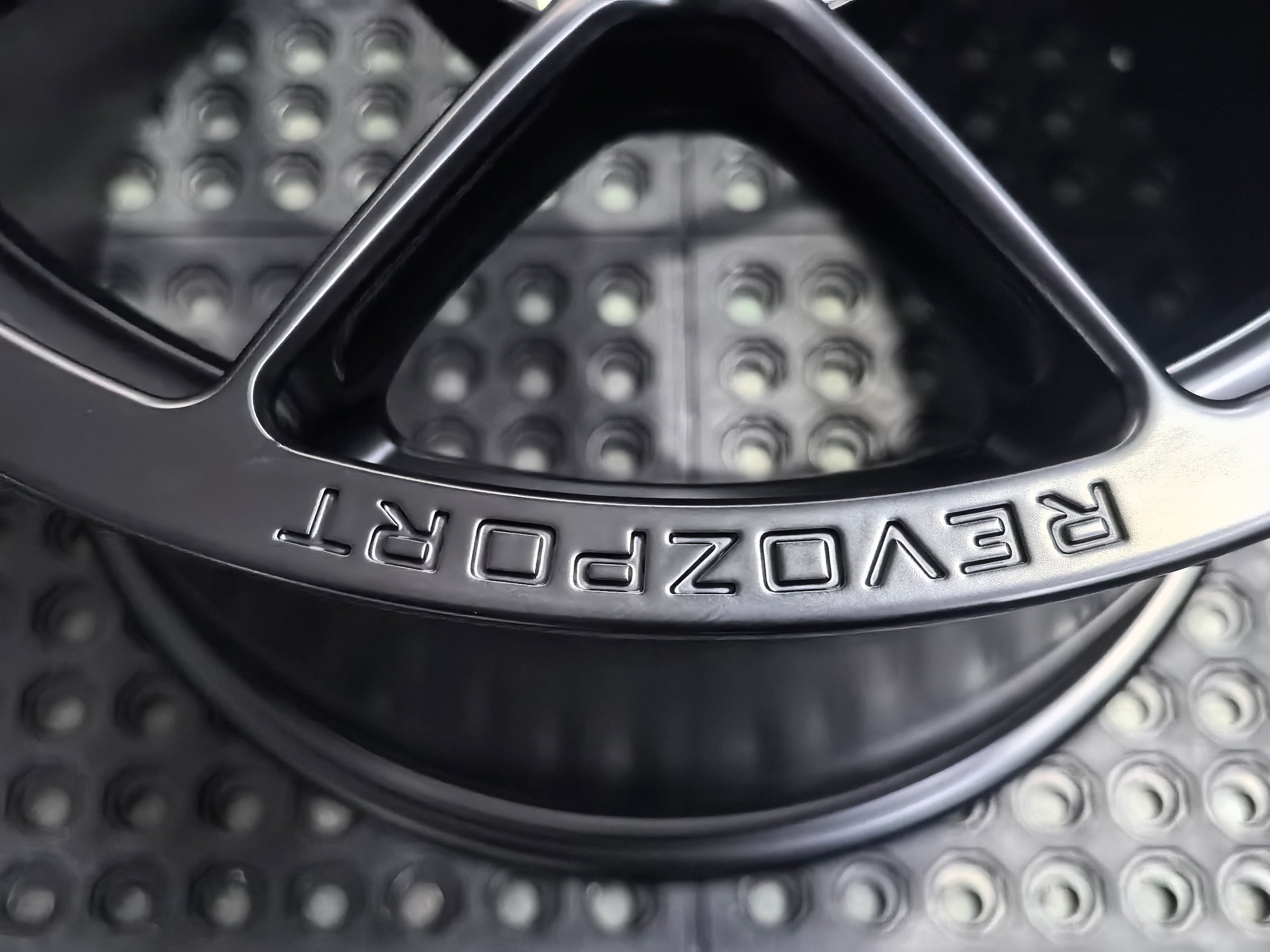 Studio-style close-up of a matte black forged performance wheel featuring a lightweight multi-spoke design and REVOZPORT branding. The wheel is displayed on a textured industrial floor, highlighting its premium finish, deep concave profile, and motorsport-inspired styling.