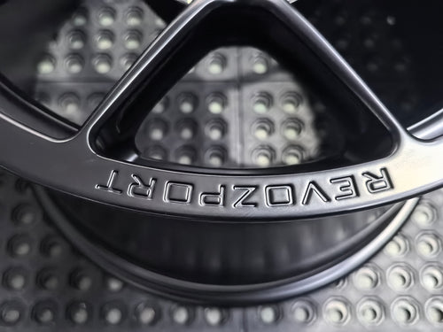 Studio-style close-up of a matte black forged performance wheel featuring a lightweight multi-spoke design and REVOZPORT branding. The wheel is displayed on a textured industrial floor, highlighting its premium finish, deep concave profile, and motorsport-inspired styling.