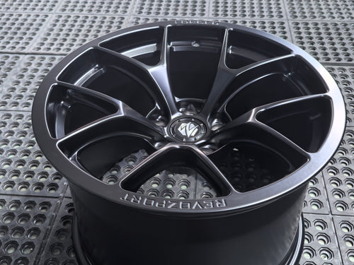 Studio-style close-up of a matte black forged performance wheel featuring a lightweight multi-spoke design and REVOZPORT branding. The wheel is displayed on a textured industrial floor, highlighting its premium finish, deep concave profile, and motorsport-inspired styling.