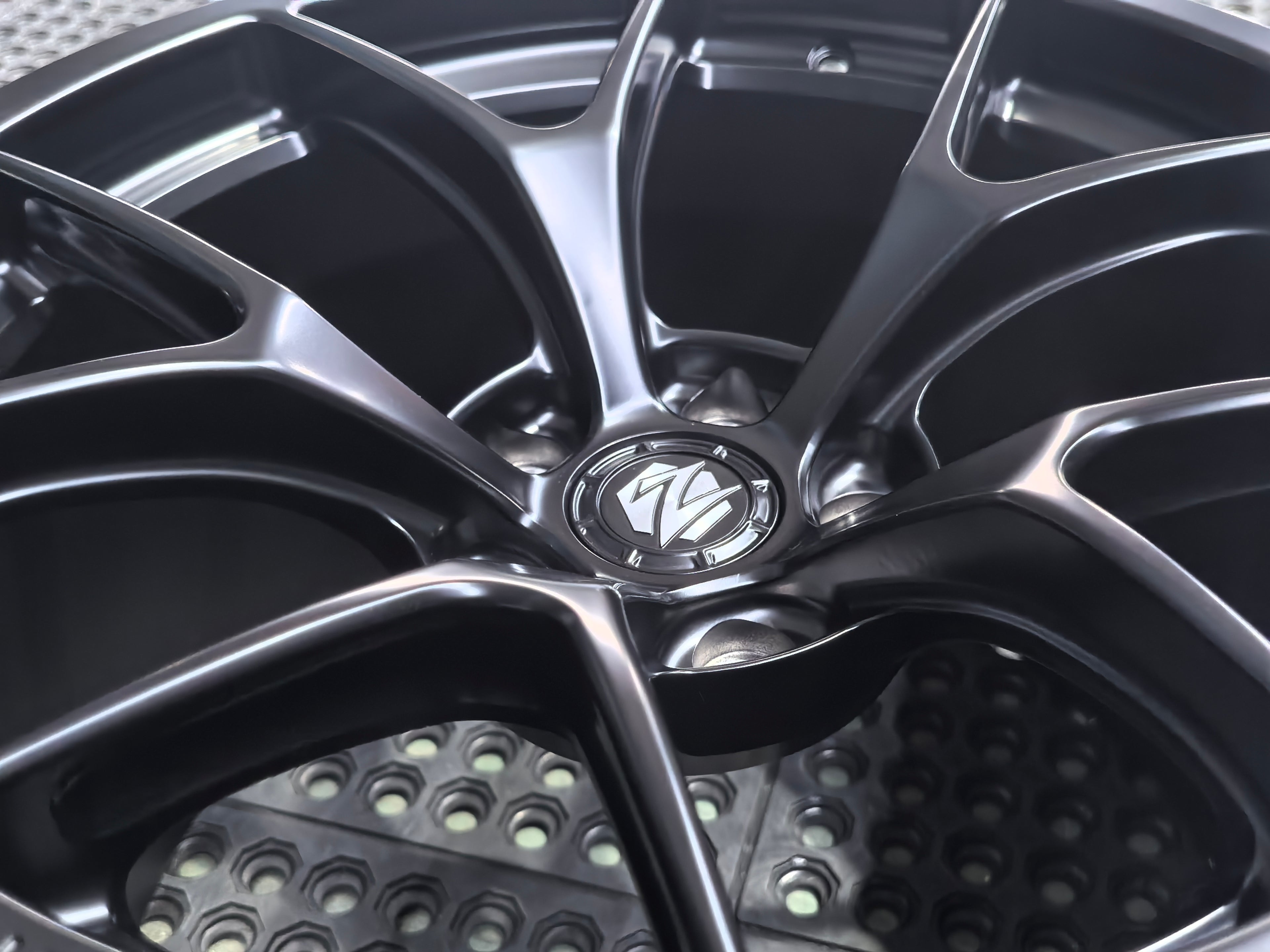 Studio-style close-up of a matte black forged performance wheel featuring a lightweight multi-spoke design and REVOZPORT branding. The wheel is displayed on a textured industrial floor, highlighting its premium finish, deep concave profile, and motorsport-inspired styling.