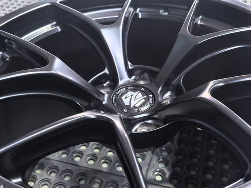 Studio-style close-up of a matte black forged performance wheel featuring a lightweight multi-spoke design and REVOZPORT branding. The wheel is displayed on a textured industrial floor, highlighting its premium finish, deep concave profile, and motorsport-inspired styling.