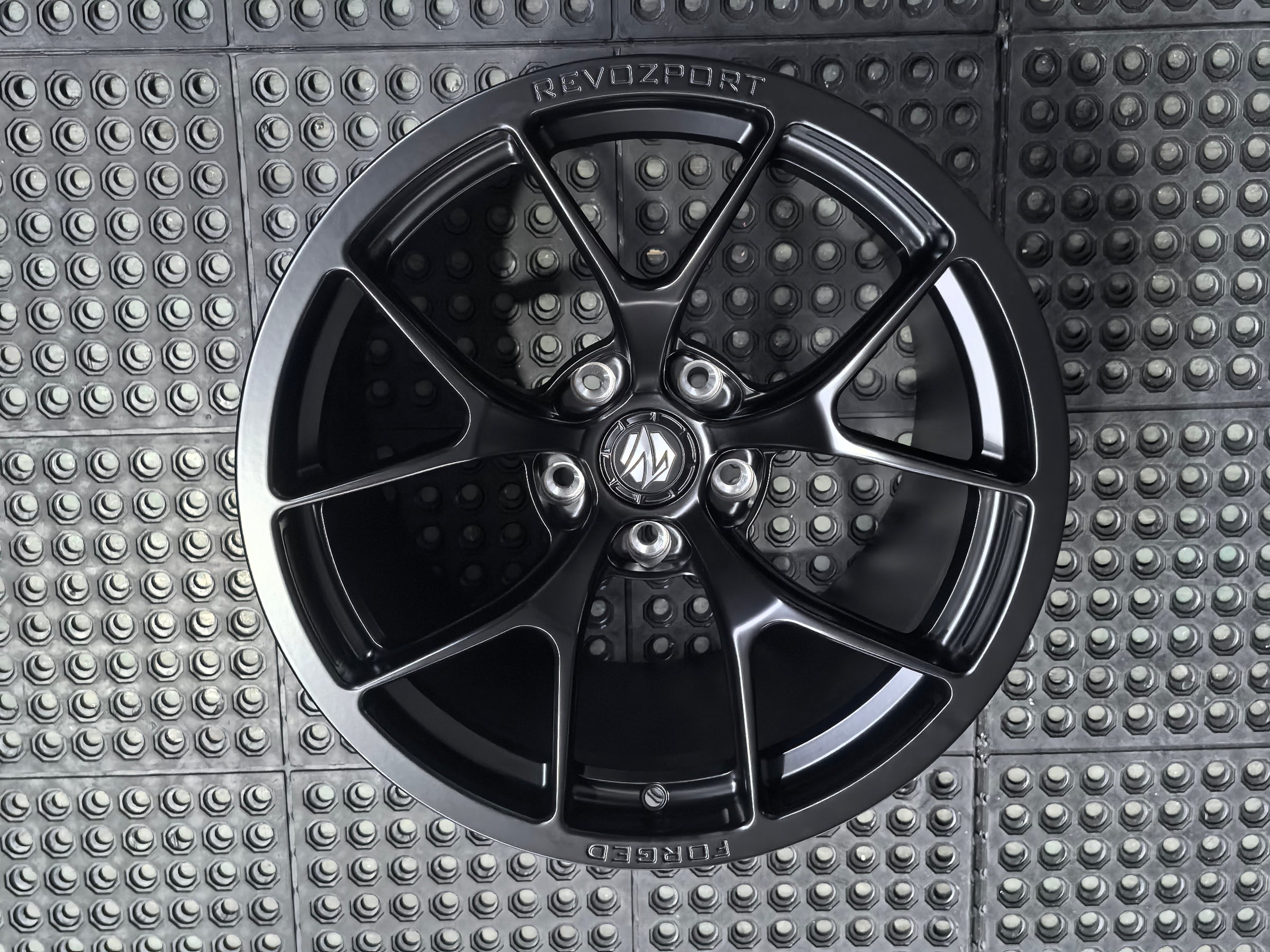 Studio-style close-up of a matte black forged performance wheel featuring a lightweight multi-spoke design and REVOZPORT branding. The wheel is displayed on a textured industrial floor, highlighting its premium finish, deep concave profile, and motorsport-inspired styling.