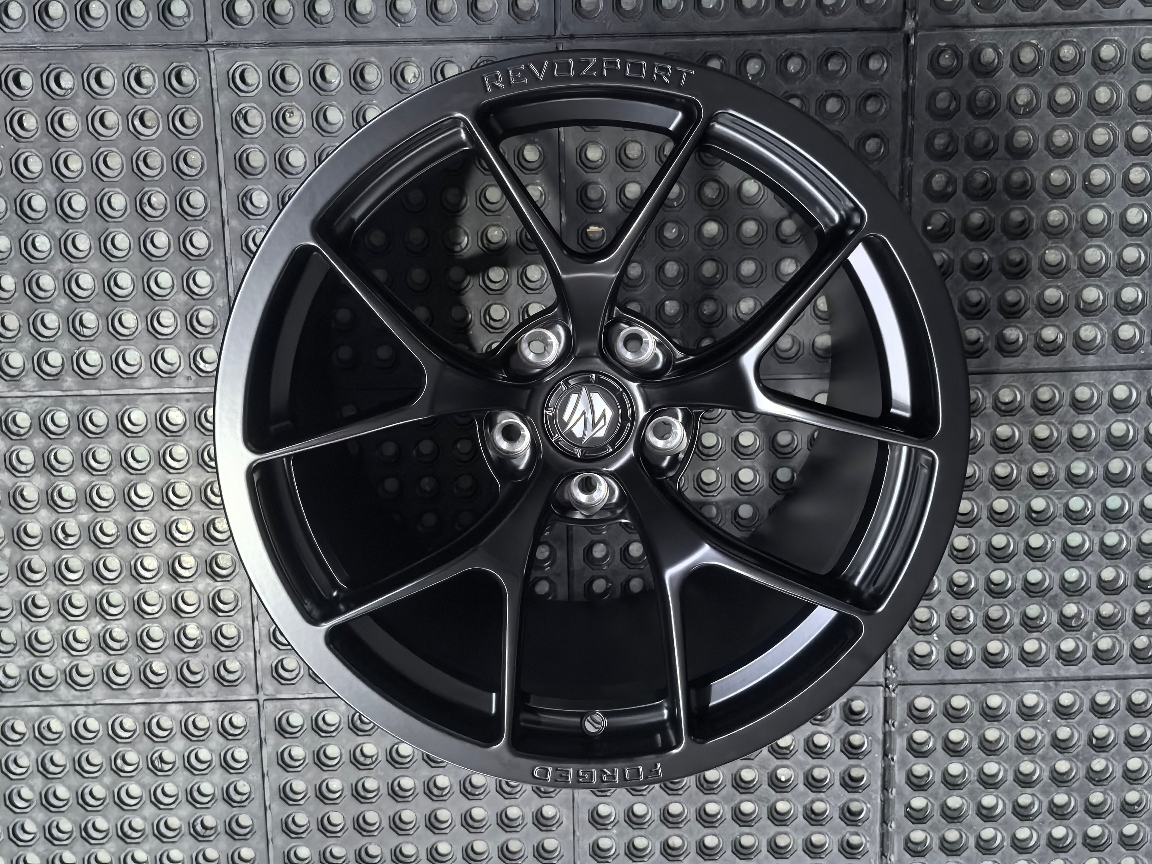 Studio-style close-up of a matte black forged performance wheel featuring a lightweight multi-spoke design and REVOZPORT branding. The wheel is displayed on a textured industrial floor, highlighting its premium finish, deep concave profile, and motorsport-inspired styling.