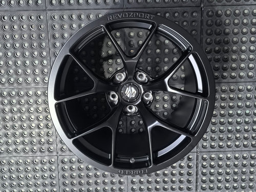 Studio-style close-up of a matte black forged performance wheel featuring a lightweight multi-spoke design and REVOZPORT branding. The wheel is displayed on a textured industrial floor, highlighting its premium finish, deep concave profile, and motorsport-inspired styling.