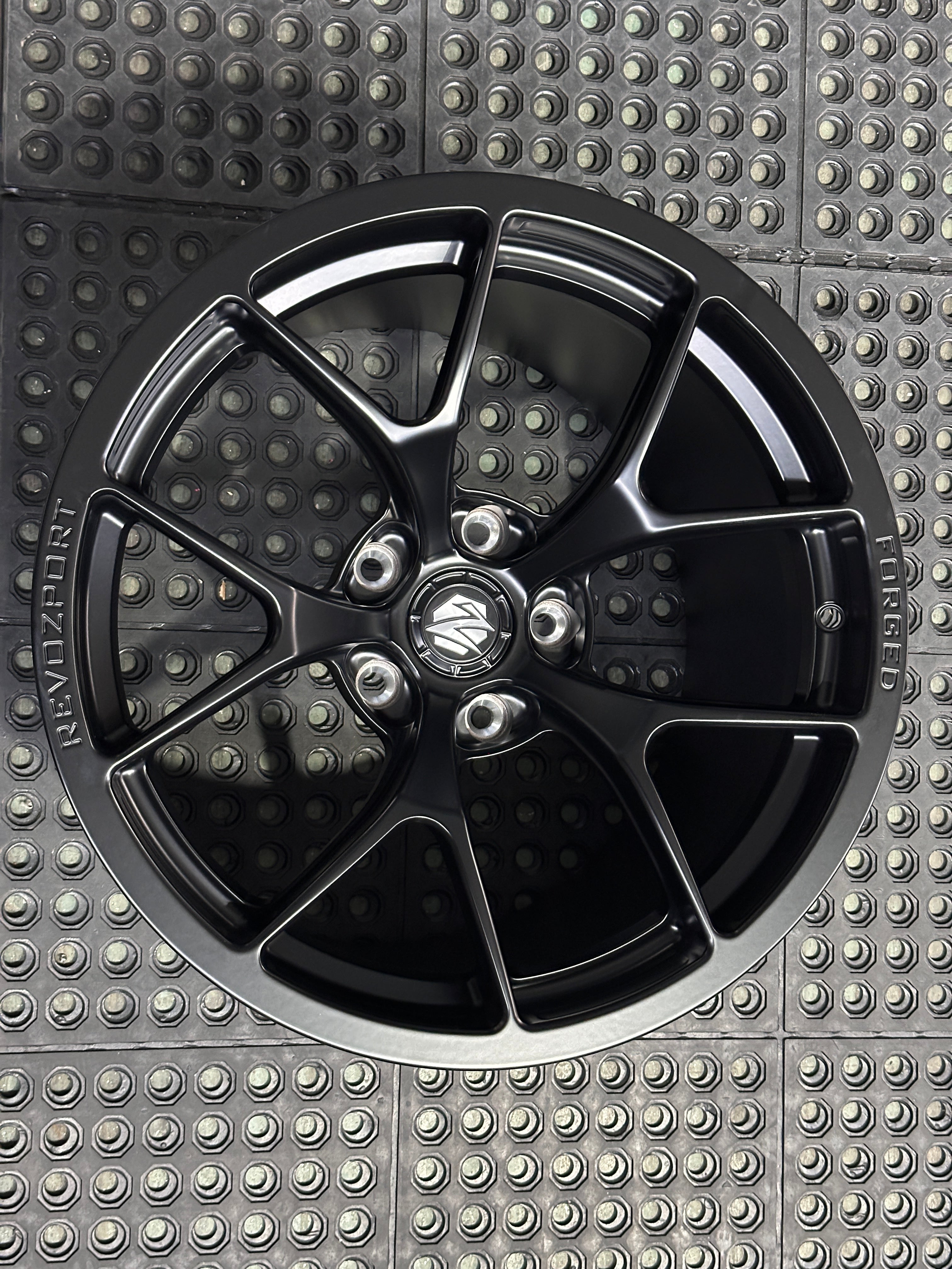 Studio-style close-up of a matte black forged performance wheel featuring a lightweight multi-spoke design and REVOZPORT branding. The wheel is displayed on a textured industrial floor, highlighting its premium finish, deep concave profile, and motorsport-inspired styling.
