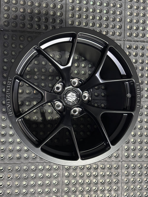 Studio-style close-up of a matte black forged performance wheel featuring a lightweight multi-spoke design and REVOZPORT branding. The wheel is displayed on a textured industrial floor, highlighting its premium finish, deep concave profile, and motorsport-inspired styling.