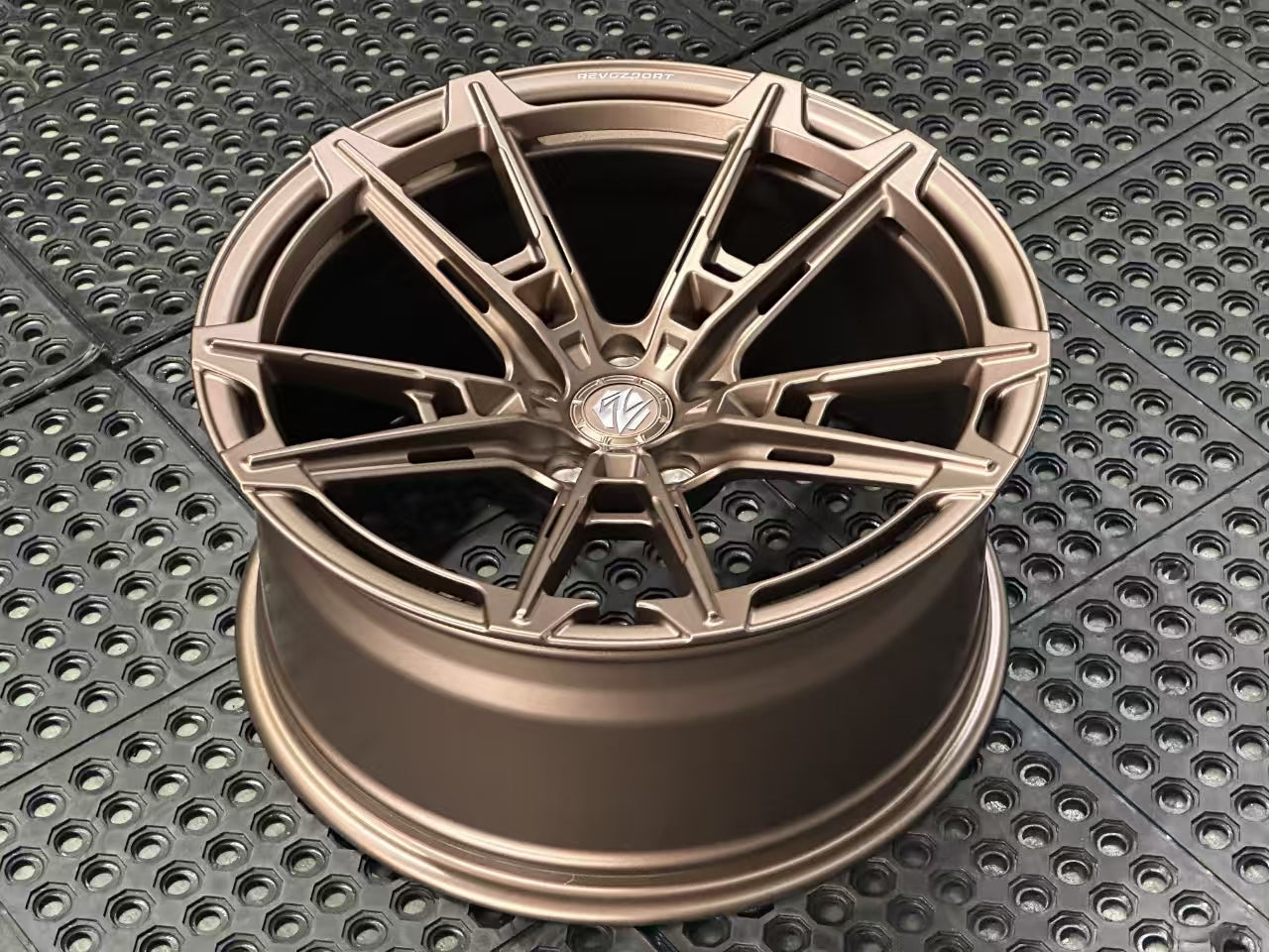 Bronze REVOZPORT forged alloy wheel with an intricate split multi-spoke design displayed on a black textured workshop floor.