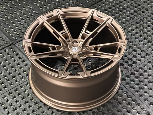 Bronze REVOZPORT forged alloy wheel with an intricate split multi-spoke design displayed on a black textured workshop floor.