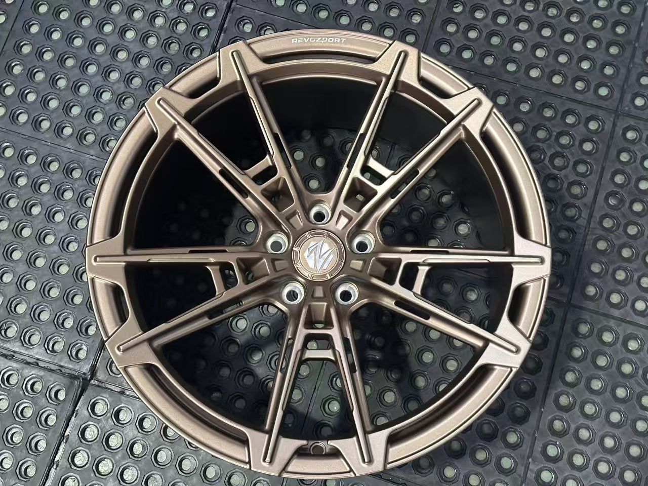 Bronze REVOZPORT forged alloy wheel with an intricate split multi-spoke design displayed on a black textured workshop floor.