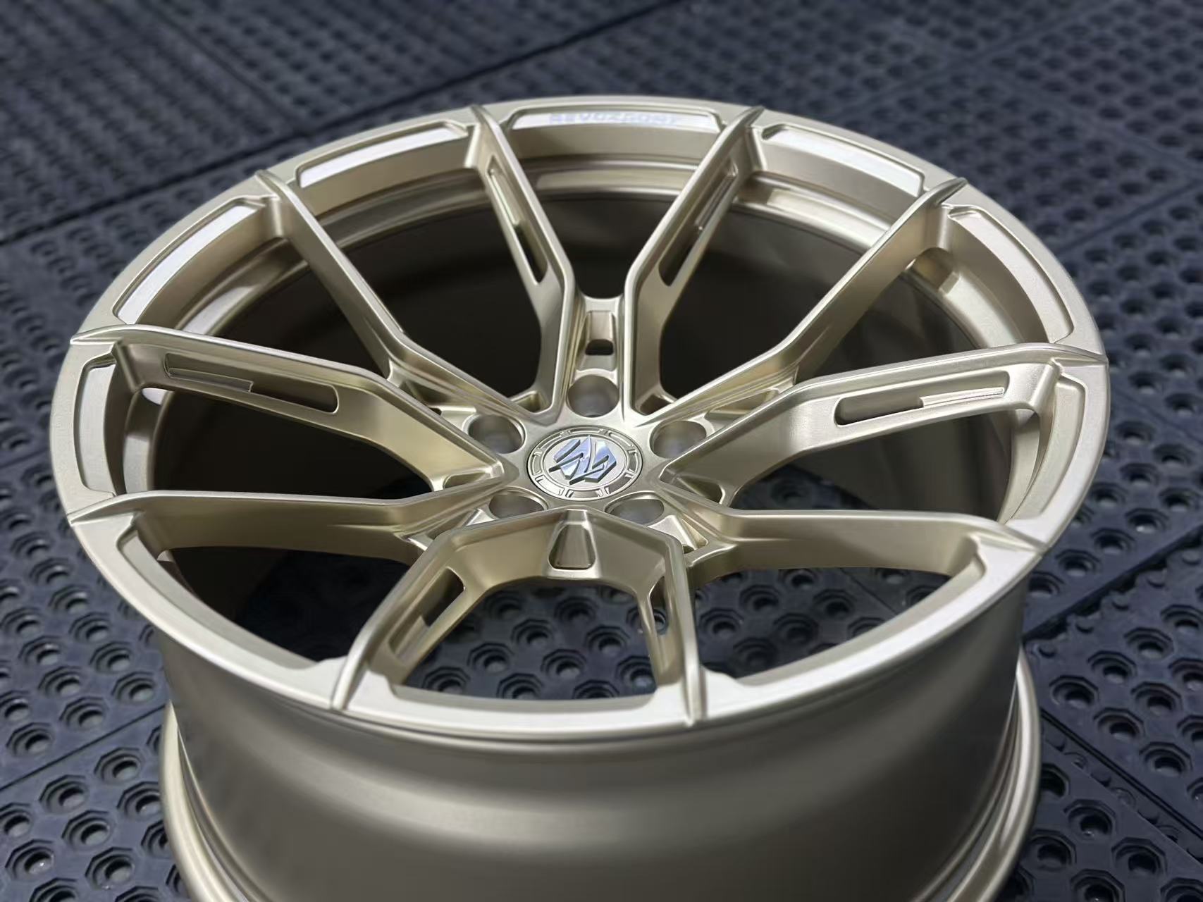 Bronze REVOZPORT forged alloy wheel with split multi-spoke design displayed on a black textured workshop floor.