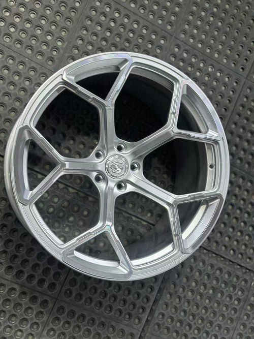Silver forged alloy wheel with a multi-spoke design displayed against a black textured rubber floor.