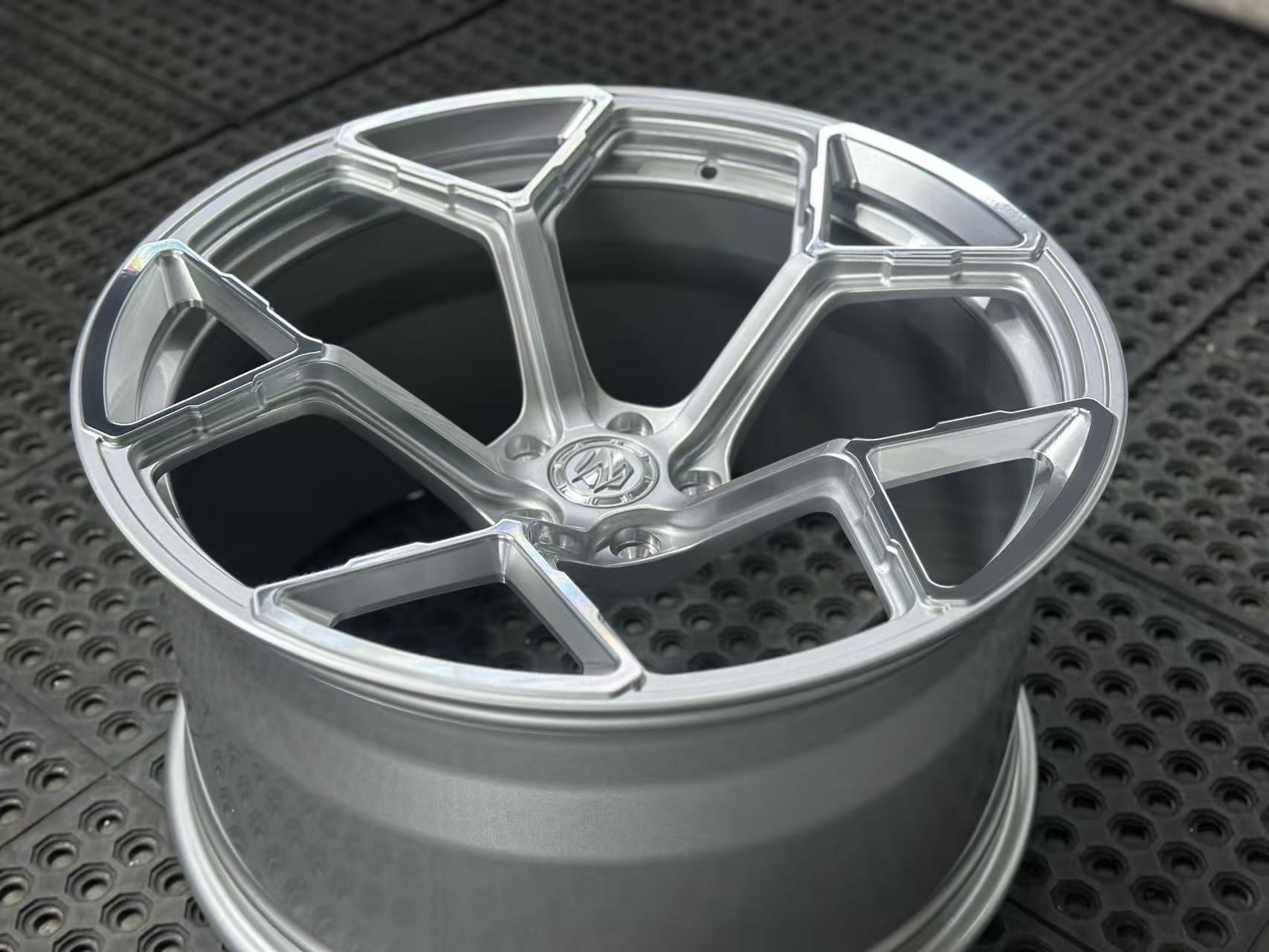 Silver forged alloy wheel with a multi-spoke design displayed against a black textured rubber floor.