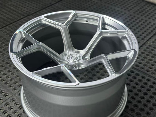 Silver forged alloy wheel with a multi-spoke design displayed against a black textured rubber floor.