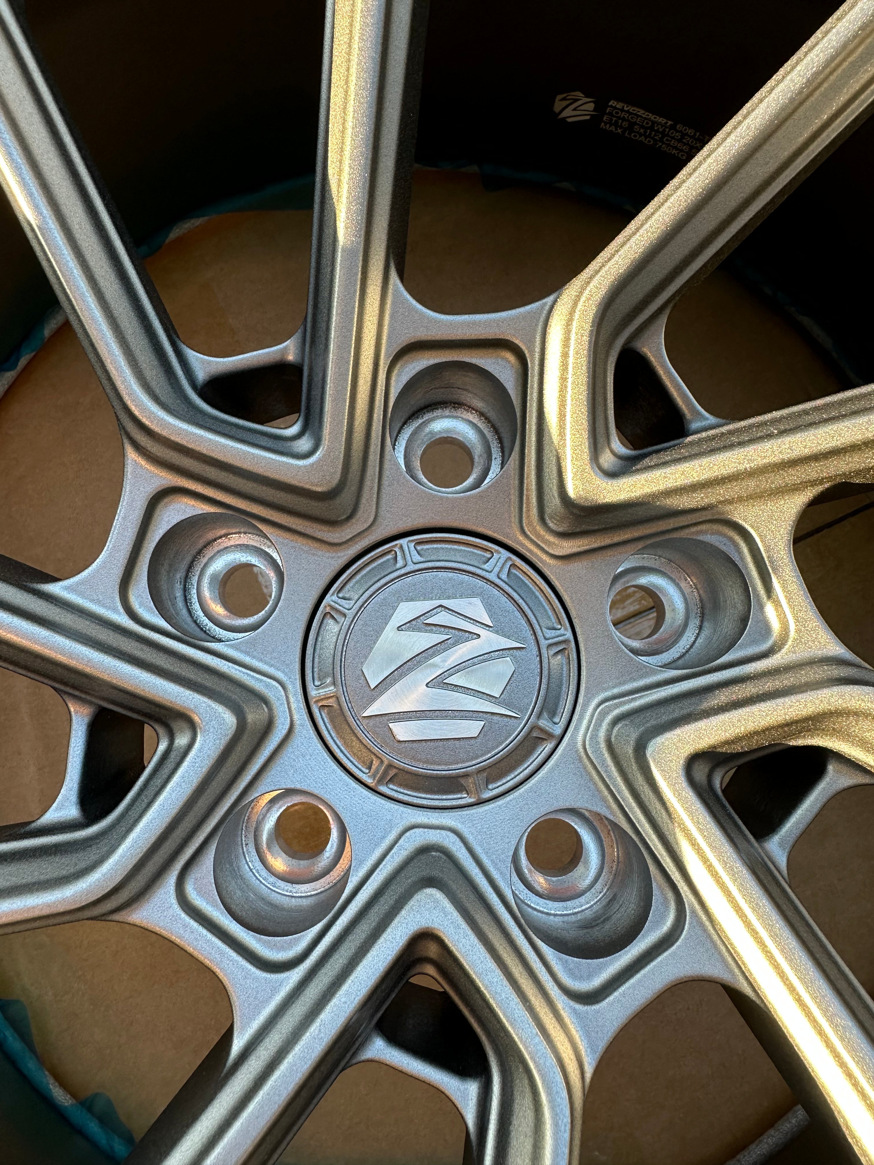 Matte gray REVOZPORT forged alloy wheel with an intricate split multi-spoke design displayed on a black textured workshop floor.
