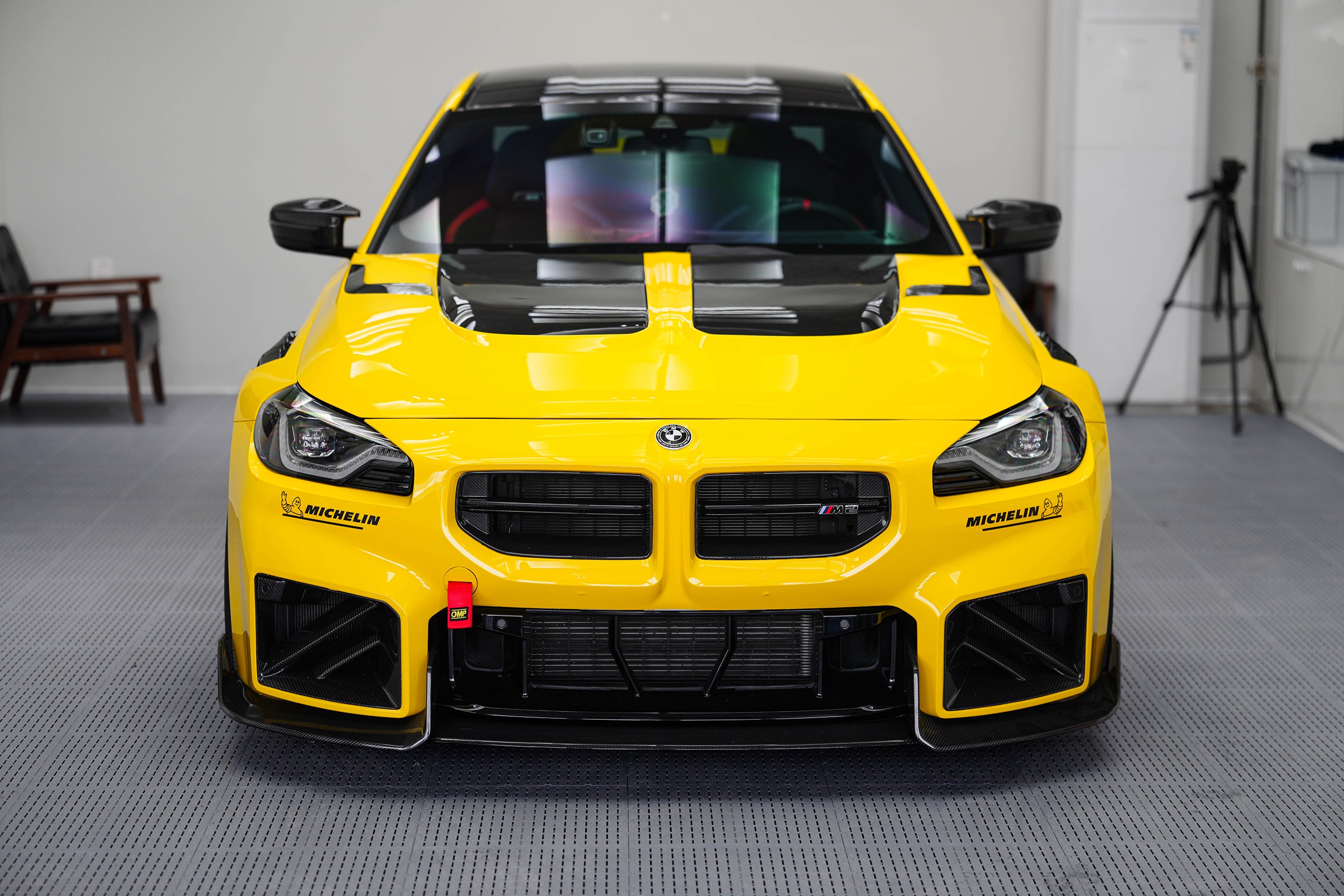 Yellow BMW G87 M2 featuring a REVOZPORT carbon fiber body kit with vented hood, front splitter, large air intakes, and aggressive track-focused aero styling displayed indoors.