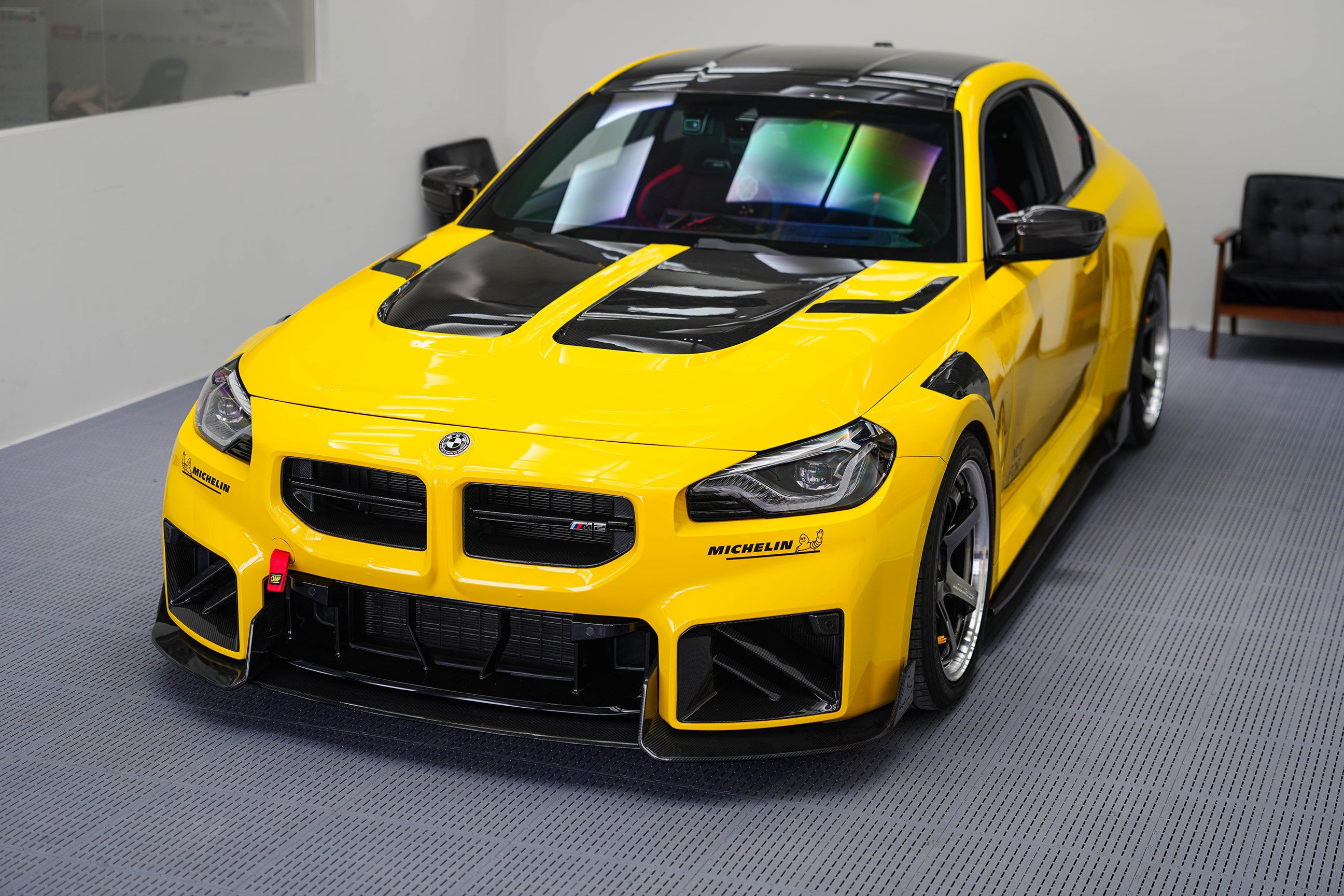 Yellow BMW G87 M2 featuring a REVOZPORT carbon fiber body kit with vented hood, front splitter, large air intakes, and aggressive track-focused aero styling displayed indoors.