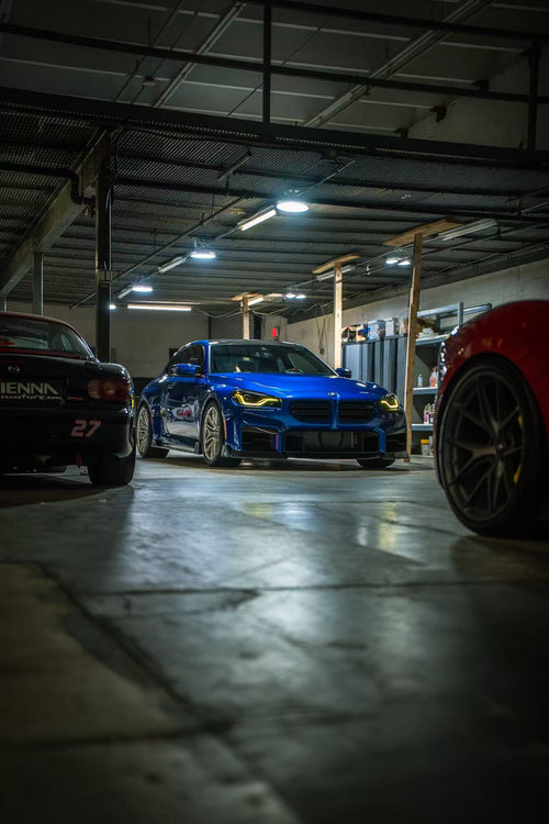 Blue BMW G87 M2 featuring a REVOZPORT carbon fiber front lip, aggressive aero styling, and yellow DRL headlights displayed inside a workshop.