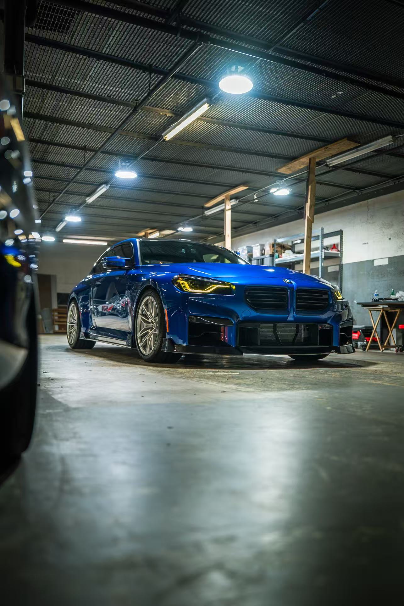 Blue BMW G87 M2 featuring a REVOZPORT carbon fiber front lip, aggressive aero styling, and yellow DRL headlights displayed inside a workshop.