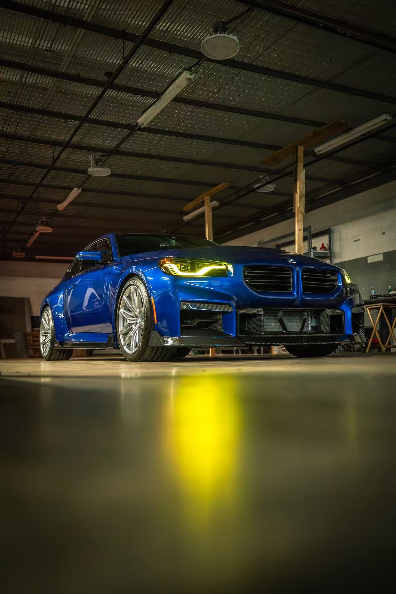 Blue BMW G87 M2 featuring a REVOZPORT carbon fiber front lip, aggressive aero styling, and yellow DRL headlights displayed inside a workshop.
