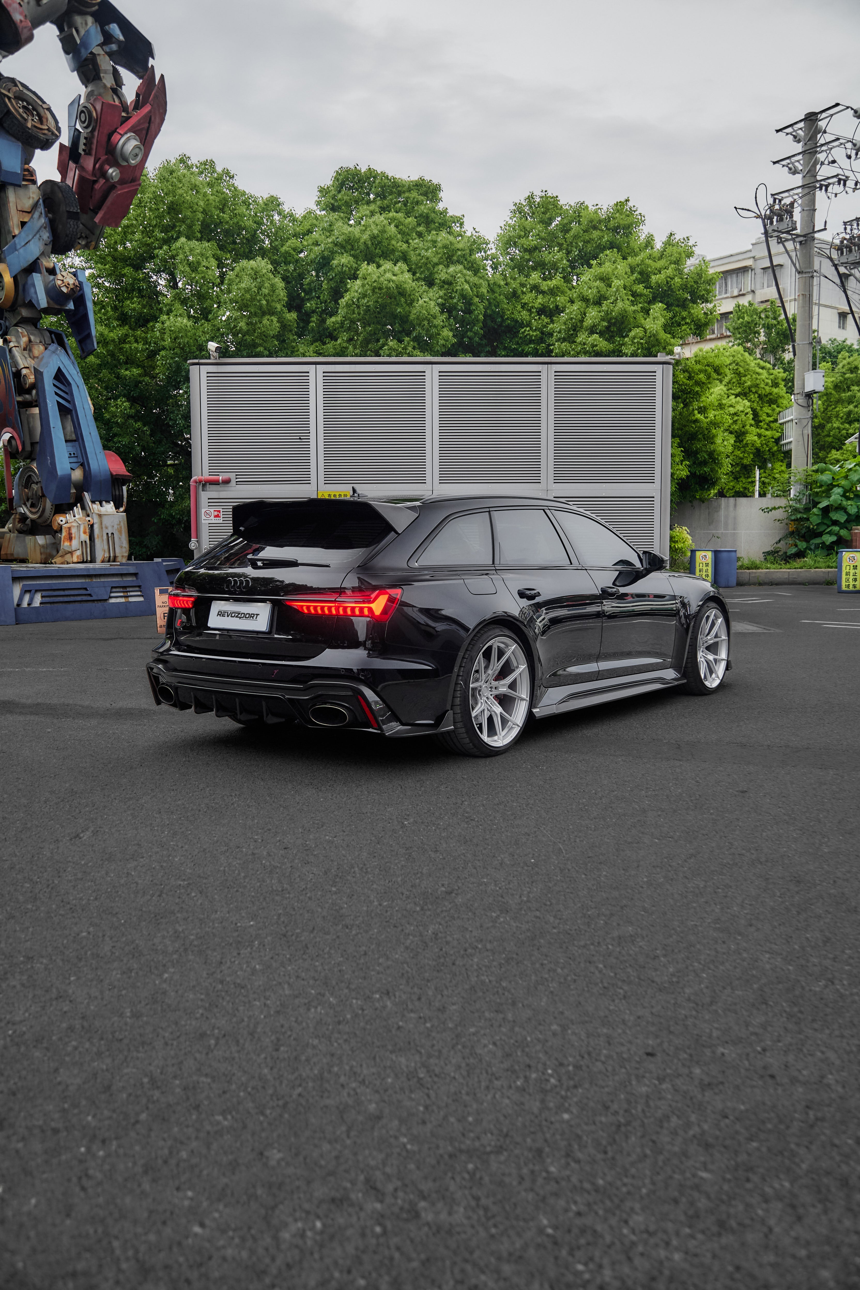 Black Audi RS6 Avant fitted with a REVOZPORT body kit and silver multi-spoke wheels, parked in an open lot surrounded by trees.
