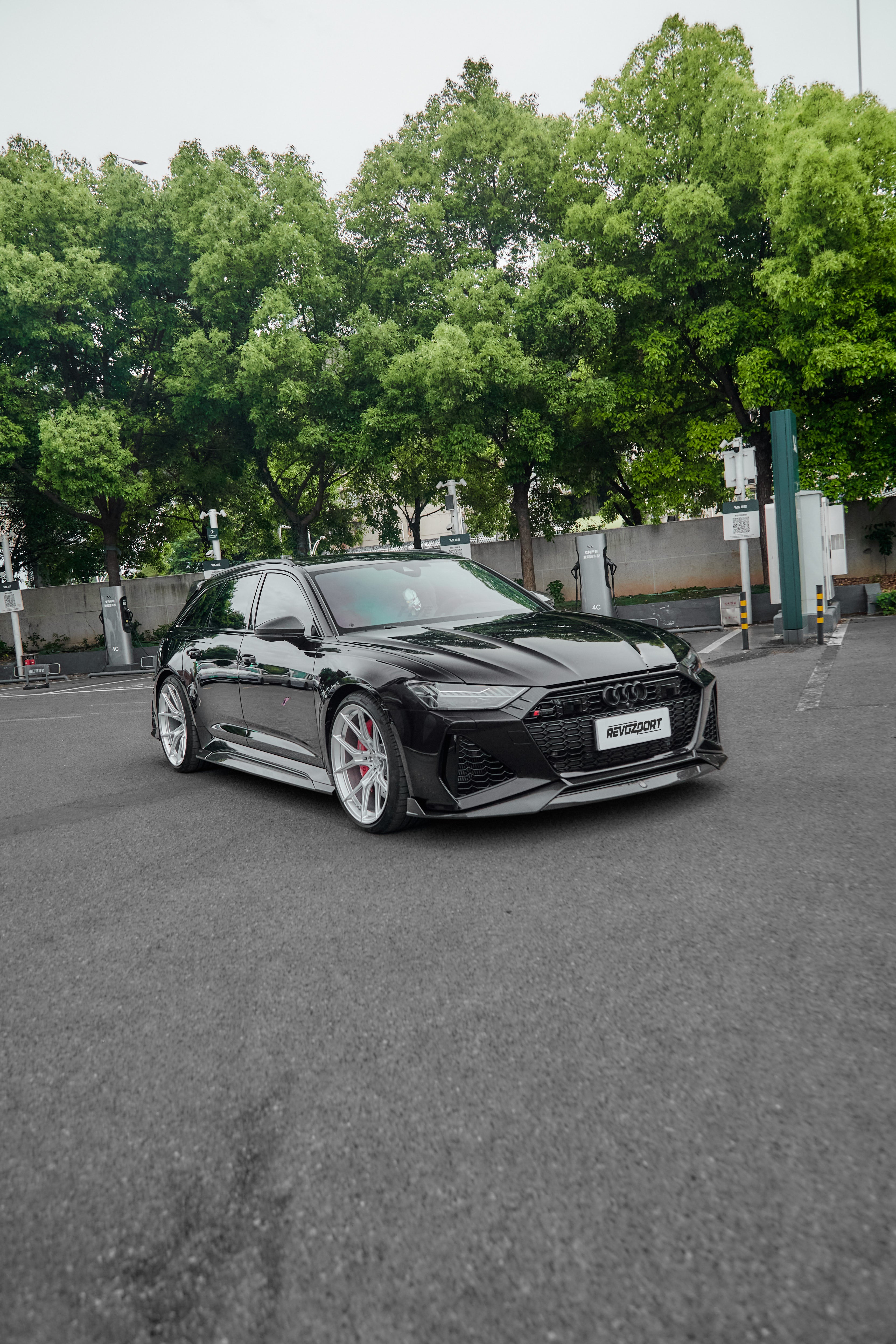 Black Audi RS6 Avant fitted with a REVOZPORT body kit and silver multi-spoke wheels, parked in an open lot surrounded by trees.