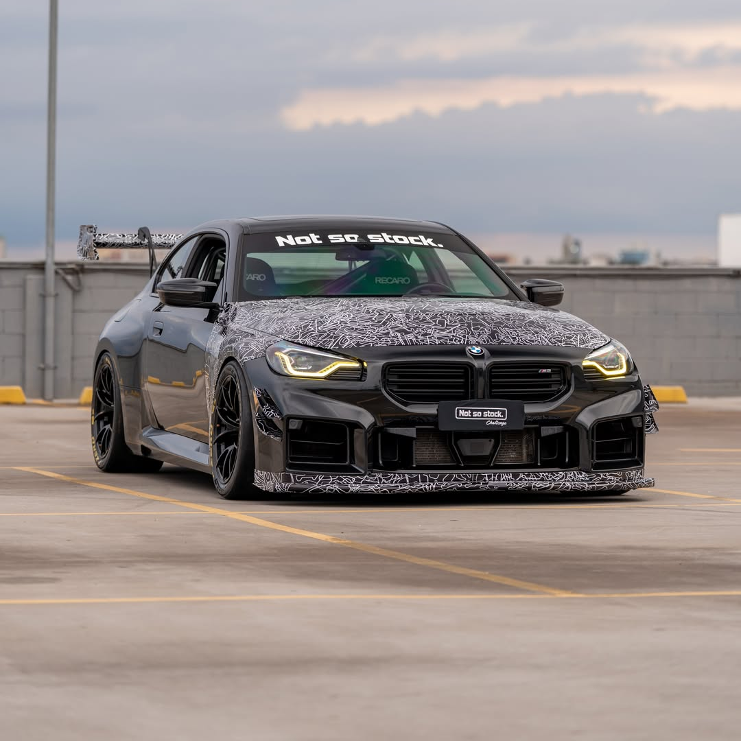 Gray BMW M2 featuring a REVOZPORT body kit with front lip, rear wing, canards, vented fenders, and hood, parked in an open rooftop lot.