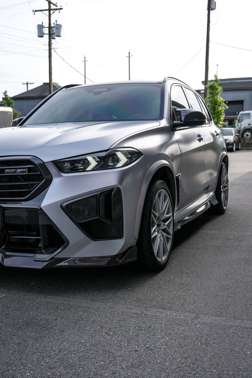 Gray BMW X5M featuring a REVOZPORT carbon fiber body kit with aggressive front lip, side skirts, and sporty aero styling parked outside a workshop.
